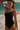 A woman is featured wearing a solid black one-piece swimsuit with a one-shoulder design and gold hardware accents on the straps. The photo is taken at a beach location with the ocean in the background.